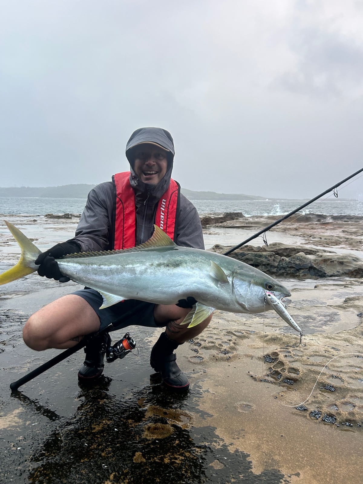 Olli-Mikael Vaittinen kneeling on a Sydney rock platform wearing a Marlin lifejacket, holding a yellowtail kingfish caught from the shore